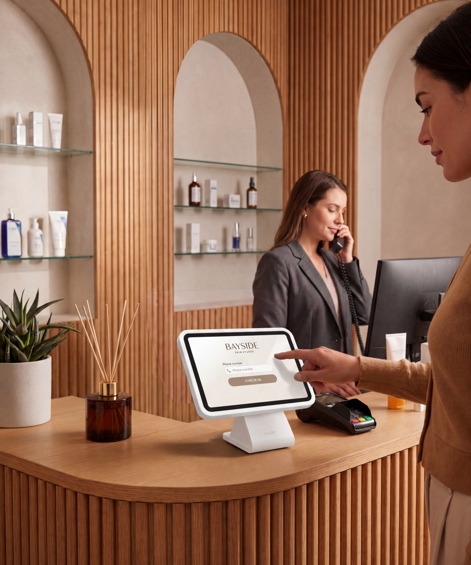 A client self-checks in on a CONNECT PRO BaseStation iPad kiosk at a warm, wood-paneled med-spa reception while a staff member takes a call — a polished check-in that matches the experience