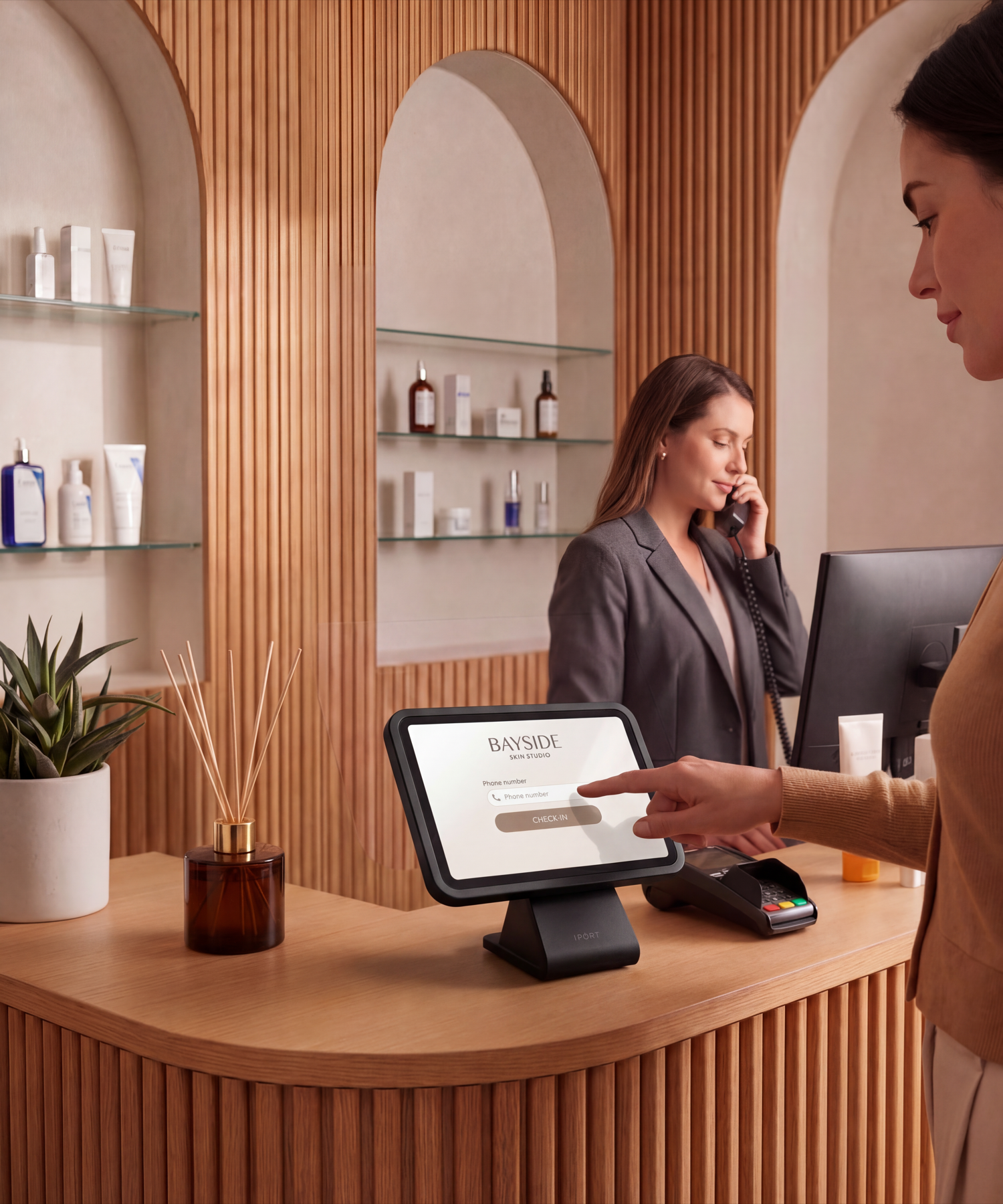 A client checks in on a black CONNECT PRO BaseStation iPad kiosk at a warm, wood-paneled medical spa front desk while a receptionist takes a call